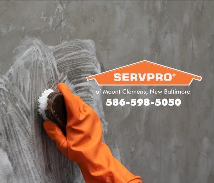 An orange-gloved hand is shown scrubbing a concrete wall with a brush. 